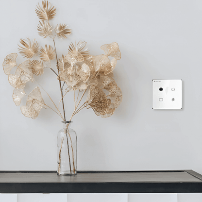 Herschel iQ Smart Scene Switch witte wandschakelaar gemonteerd op muur naast tafel met glazen vaas en decoratieve bladeren
